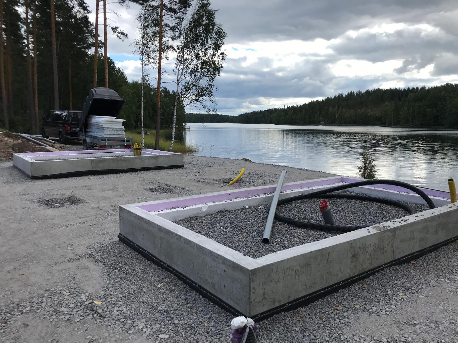 Kaksi betonista pohjaa, joissa on vaaleanpunainen eristys, täytettynä harmaalla soralla, järven rannalla metsän reunassa pilvisen taivaan alla.