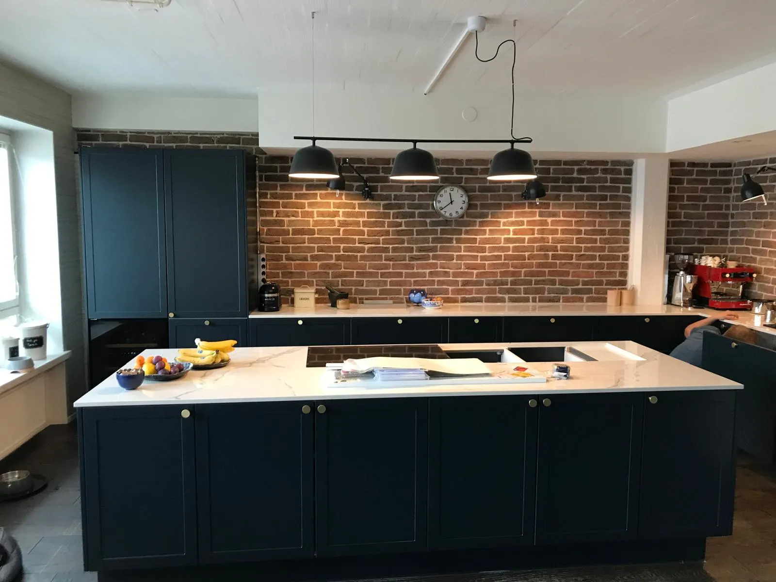 Moderni keittiö, jossa on tummansiniset kaapit, marmorinen työtaso ja tiiliseinä, valaistu kolmella mustalla riippuvalaisimella.