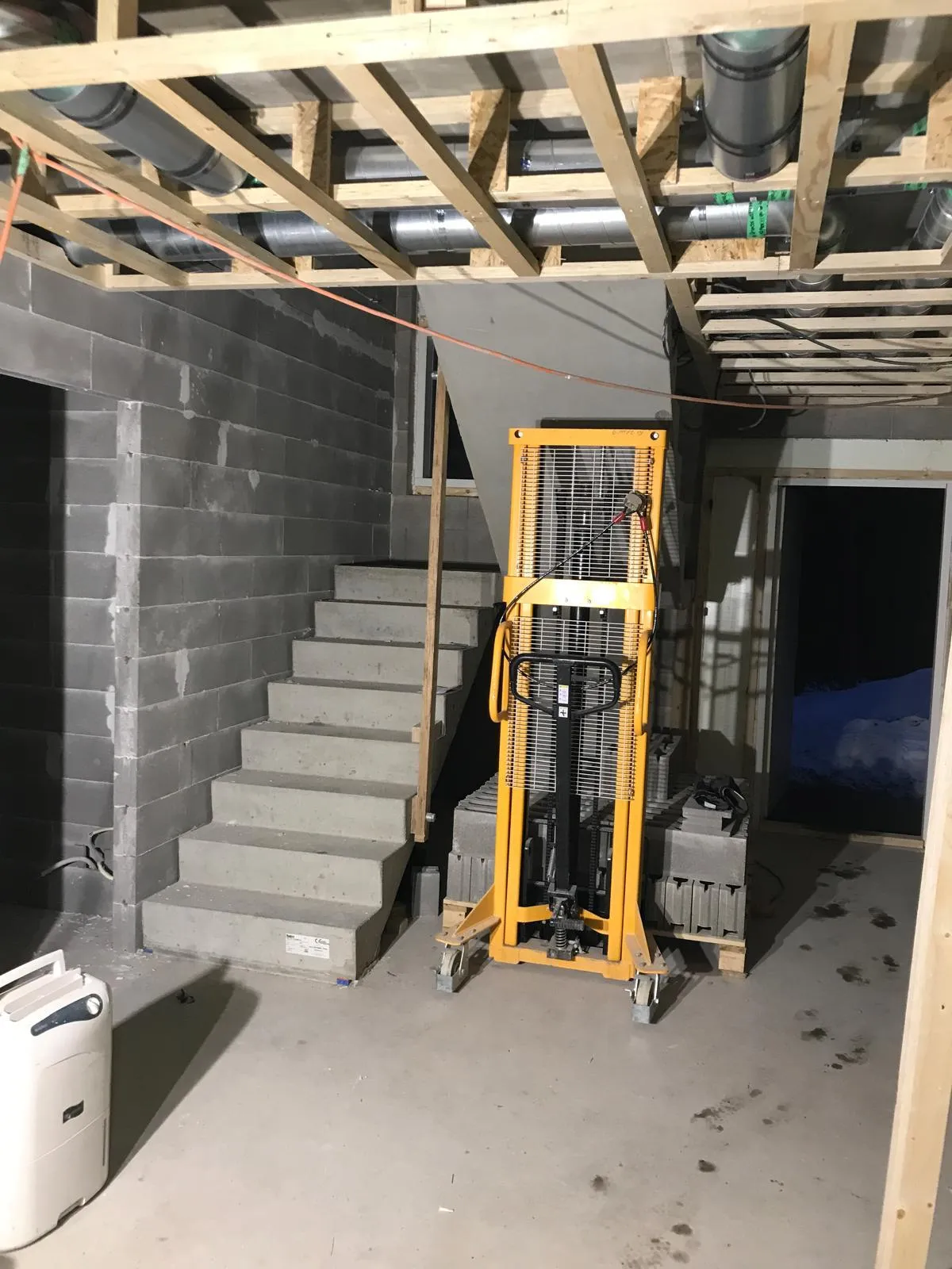 Kellaritilassa on portaat, keltainen nostolaite, ilmastointiputket katossa ja valkoinen ilmankuivain.