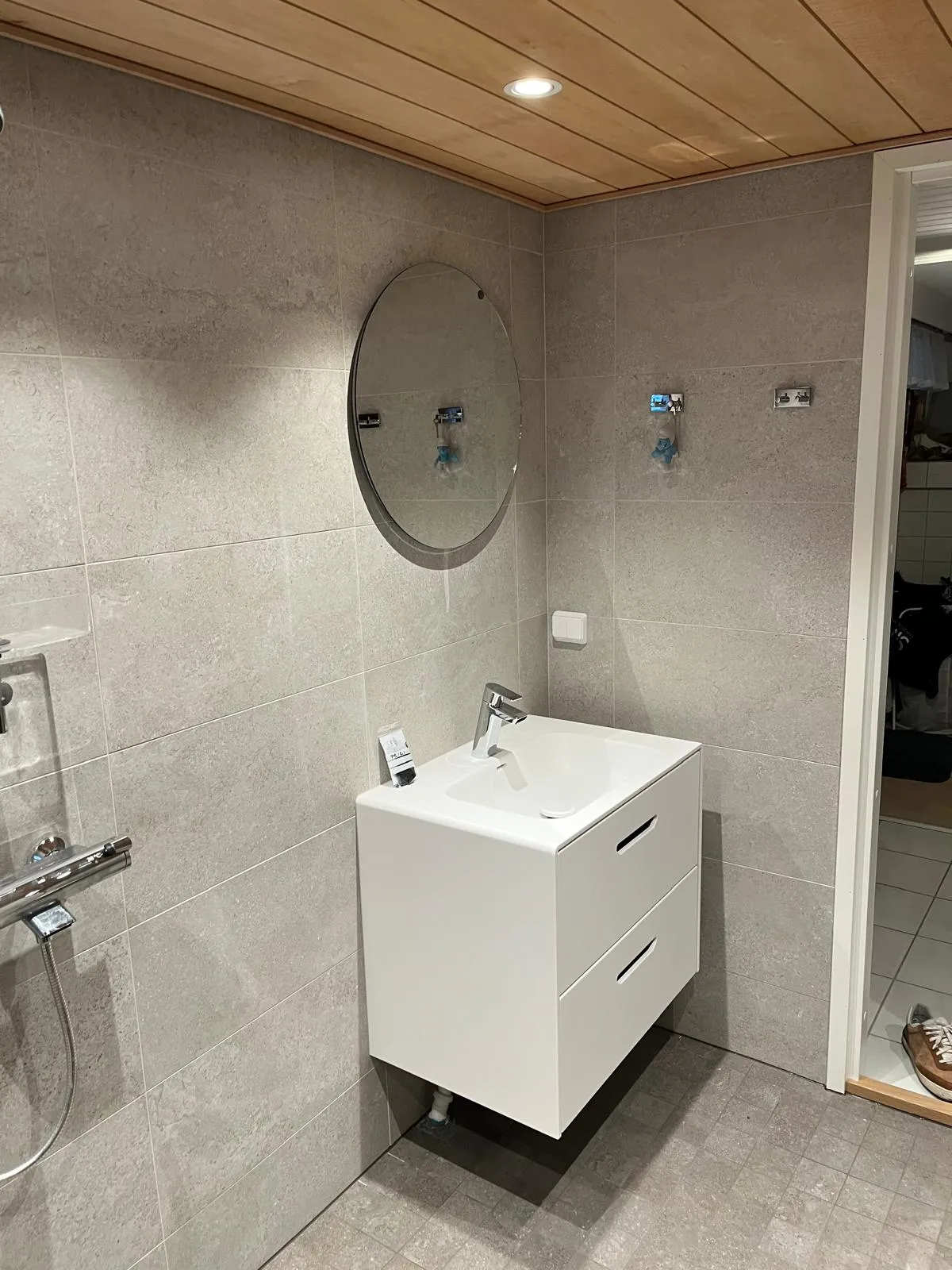 Kylpyhuoneen sisustus, jossa on pyöreä peili, valkoinen pesuallas, harmaat laatat seinillä ja puinen katto.