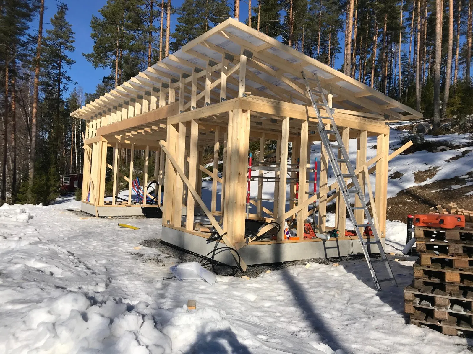 Keskeneräinen puurakenteinen rakennus lumisessa maisemassa, jossa näkyy tikkaat ja puupaletteja.