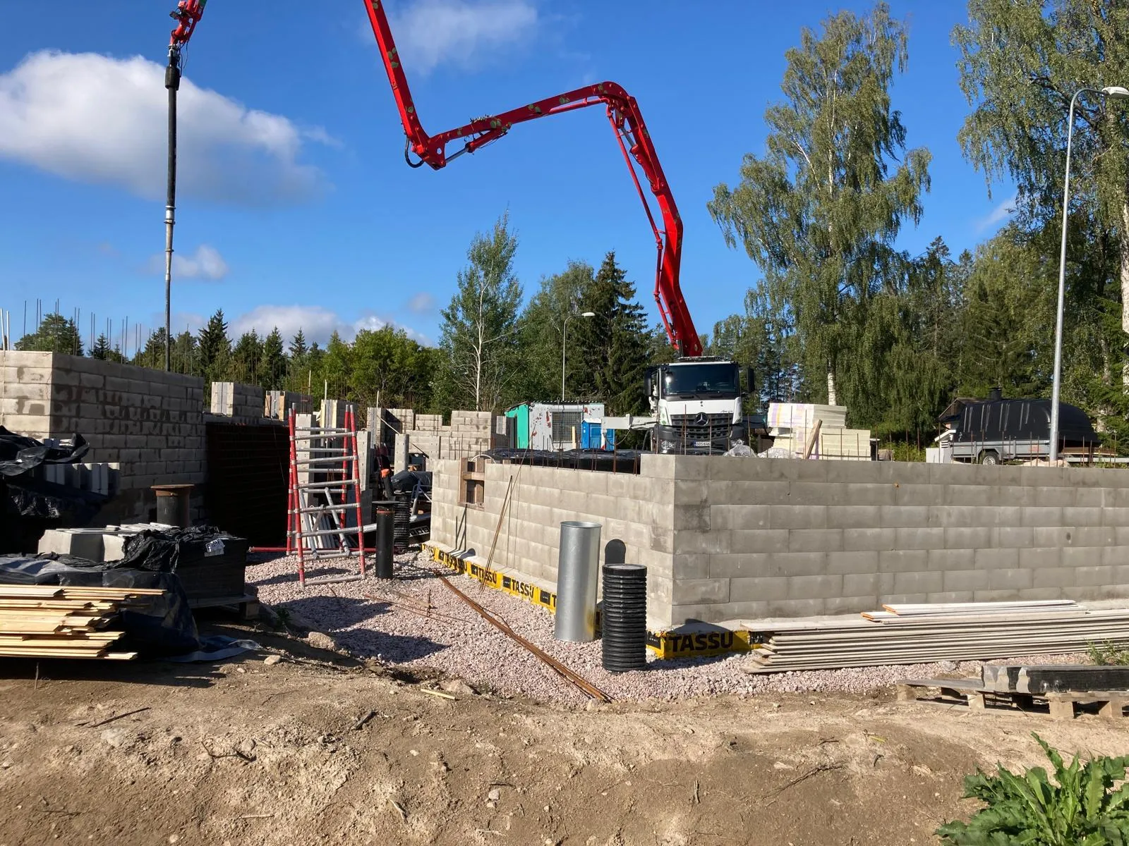 Rakennustyömaa, jossa on harmaa betoniharkkoperustus, punainen betonipumppuauto ja sininen taivas.