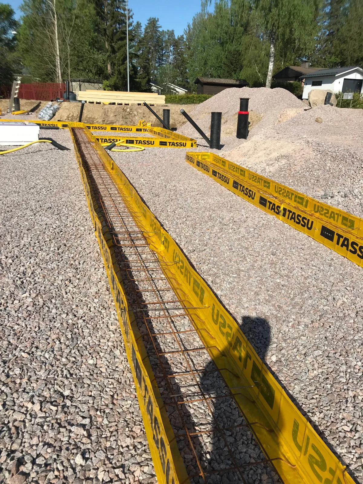 Rakennustyömaa, jossa näkyy keltainen muotti raudoituksella ja kivimurskeella, aurinkoisessa säässä.