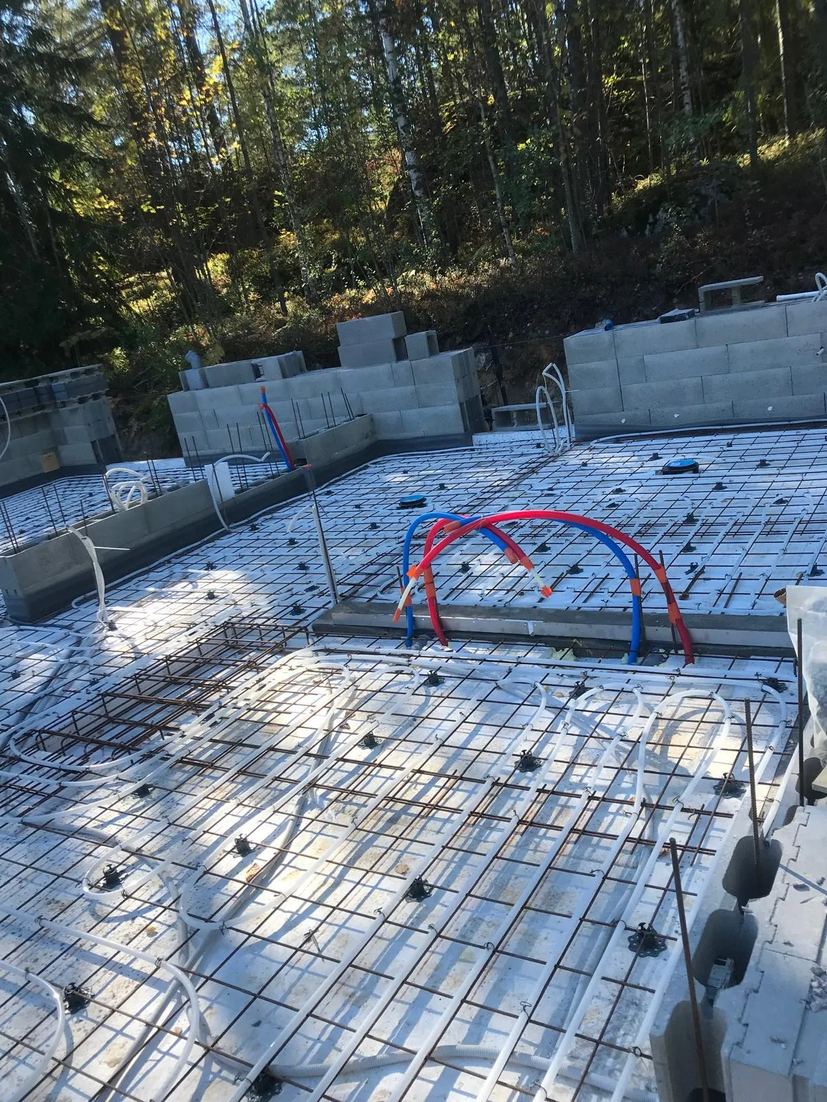 Rakennustyömaa, jossa näkyy lattialämmitysputkia, teräsverkko ja harkkoja metsäisessä ympäristössä kirkkaassa päivänvalossa.