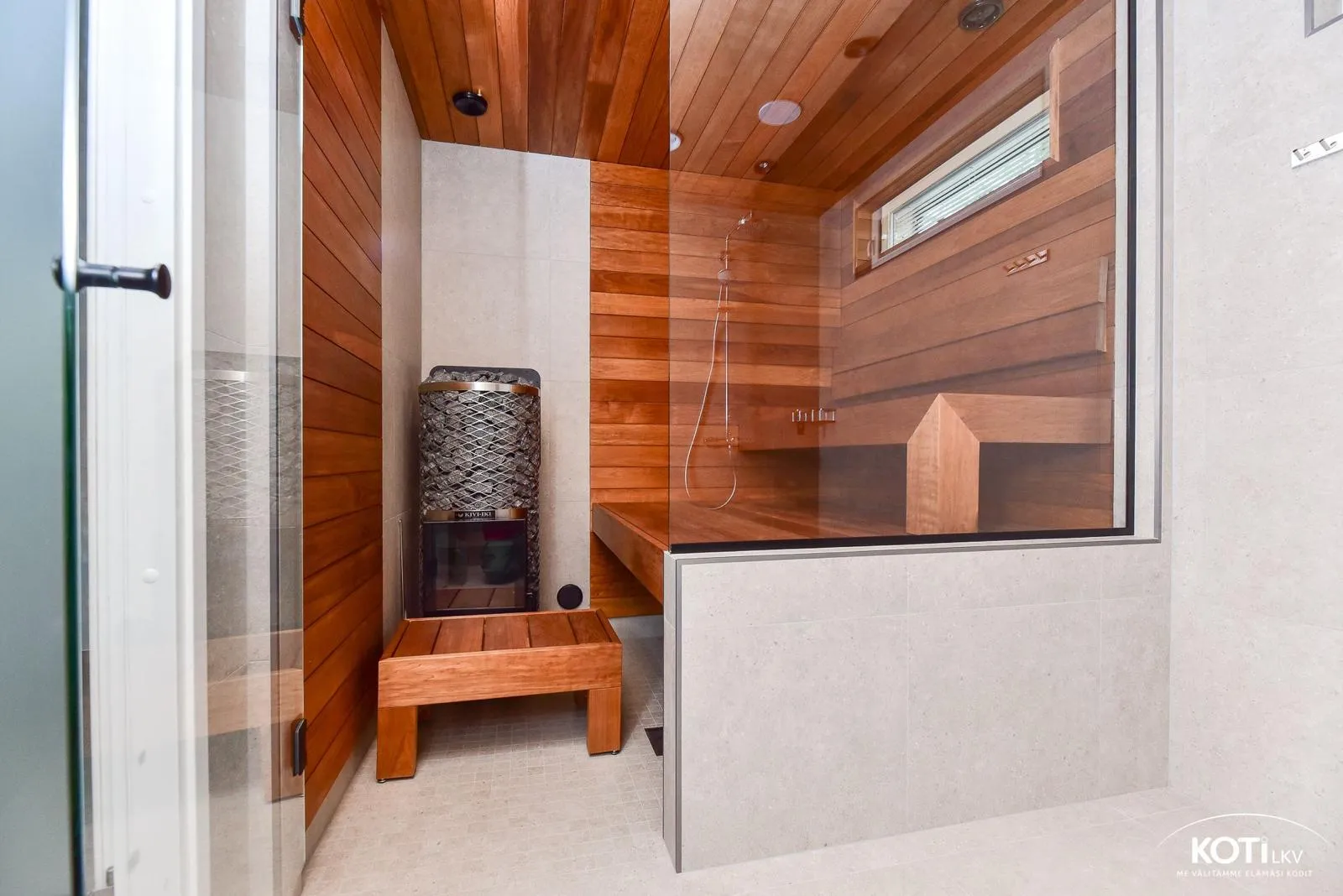 Saunan sisätila, jossa on puupanelointi, lasiseinä ja kivikiuas, valaistus on lämmin ja tunnelmallinen.