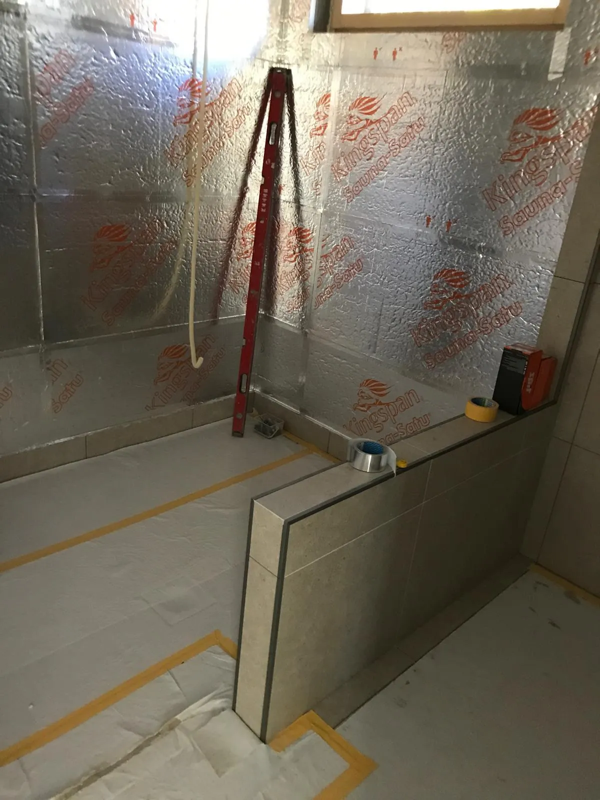 Kuva esittää rakenteilla olevaa saunaa, jossa on eristys seinissä ja lattialla. Näkyvissä on myös mittanauha, teippiä ja muita rakennustarvikkeita. Valo on kirkas ja tasainen.